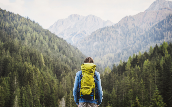 A person wearing a blue jacket and carrying a bright yellow backpack stands facing a mountain landscape.